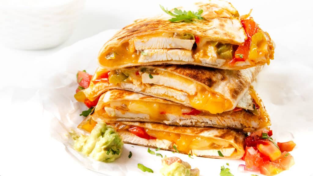 A stack of savory Chicken and Cheese Quesadillas filled with grilled chicken, melted cheese, red and green peppers, topped with fresh cilantro. Sides of guacamole and salsa are placed nearby on a white surface.