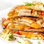 A stack of savory Chicken and Cheese Quesadillas filled with grilled chicken, melted cheese, red and green peppers, topped with fresh cilantro. Sides of guacamole and salsa are placed nearby on a white surface.