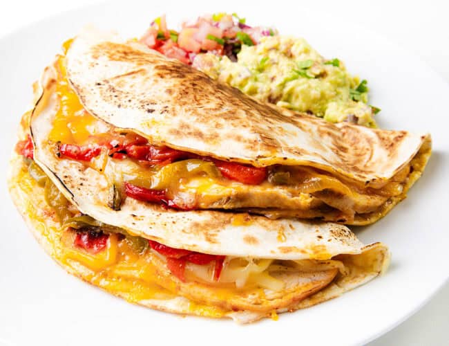 Two folded chicken and cheese quesadillas filled with melted cheese, grilled chicken, and vegetables are on a white plate. Sides of guacamole and pico de gallo accompany the dish.