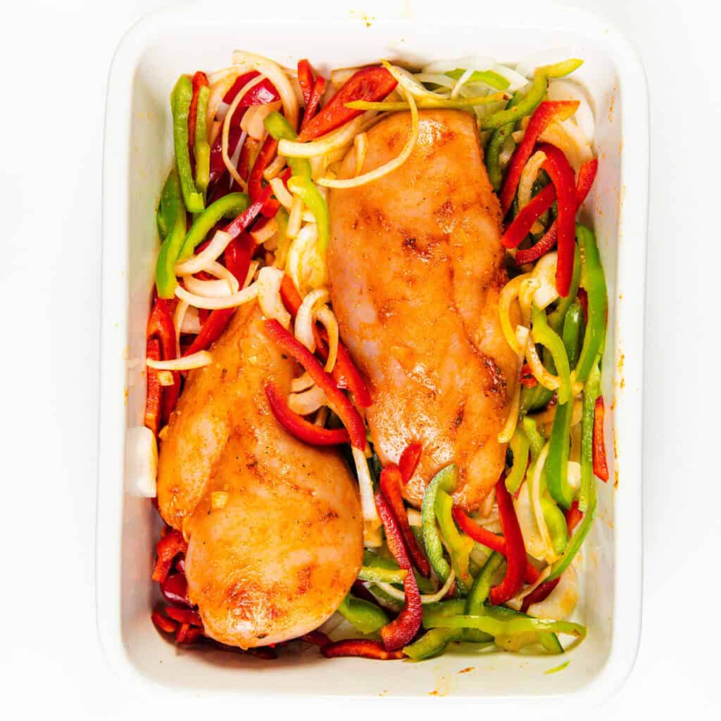 A baking dish holds two seasoned chicken breasts surrounded by sliced red, green, and orange bell peppers and onion strips, ready to be transformed into cheesy quesadillas.