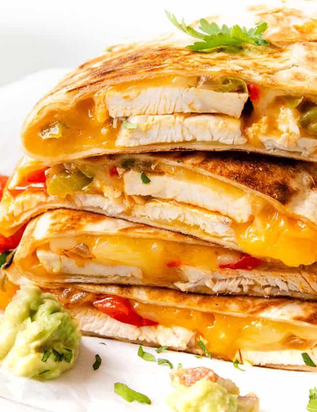 A stack of sliced quesadillas filled with melted cheese, chicken, and colorful bell peppers, garnished with cilantro. Accompanied by dollops of guacamole and diced tomatoes on a white surface.