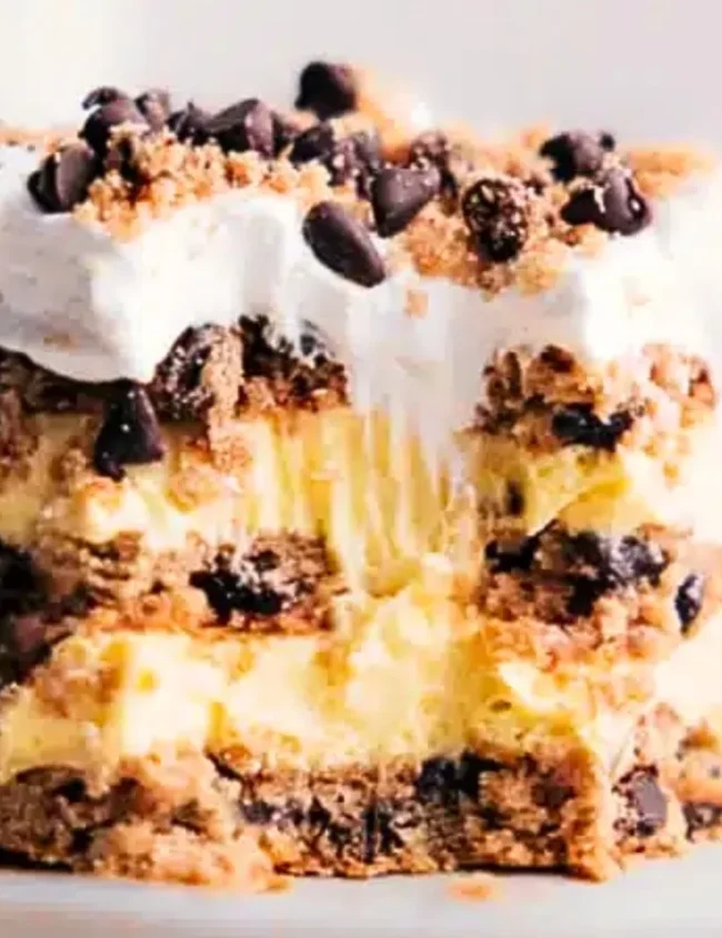 A close-up of a dessert slice with layers of cream and crushed cookies, topped with whipped cream from our favorite recipes and chocolate chips. A bite is missing from the front, and a fork rests beside it on the plate.