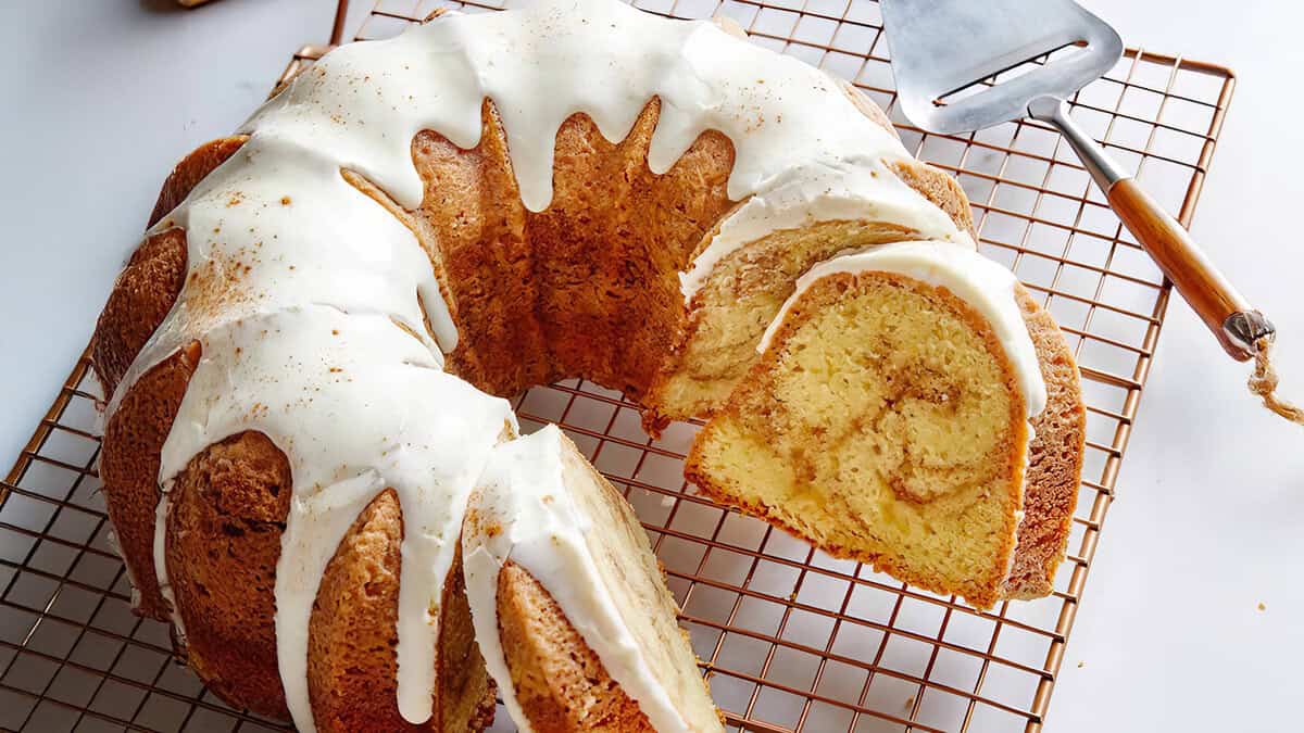 A sliced bundt cake on a cooling rack, crowned with white icing cascading down the sides. Nearby, a cake server with a wooden handle awaits. The cut reveals the cake's moist texture, embodying the essence of classic bundt cake recipes.