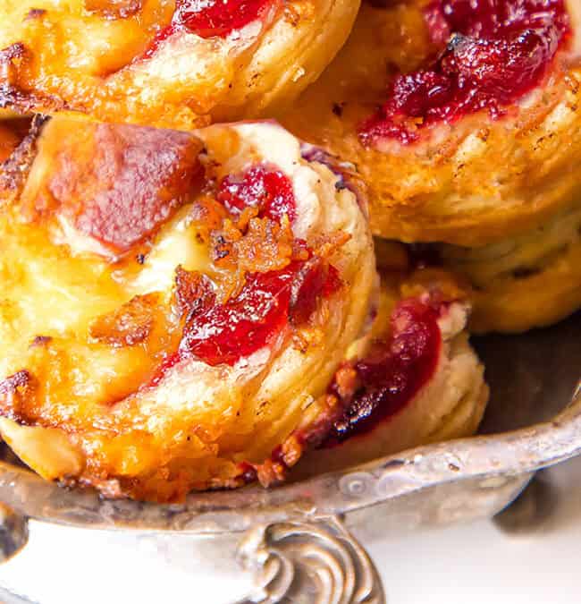 A close-up of a silver tray filled with puff pastry tarts. The tarts are topped with melted cheese, crispy bacon, and a red fruit preserve. The pastries are golden brown and slightly flaky, highlighting their freshly baked texture.