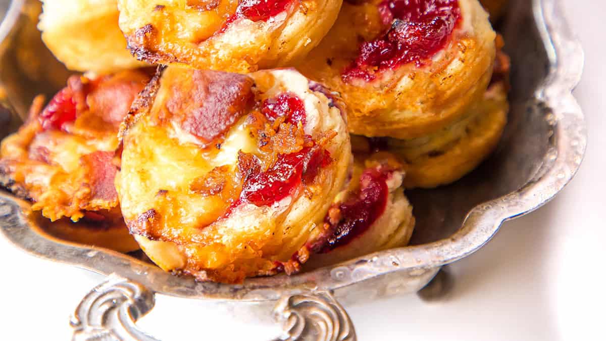 A close-up of a silver tray filled with puff pastry tarts. The tarts are topped with melted cheese, crispy bacon, and a red fruit preserve. The pastries are golden brown and slightly flaky, highlighting their freshly baked texture.