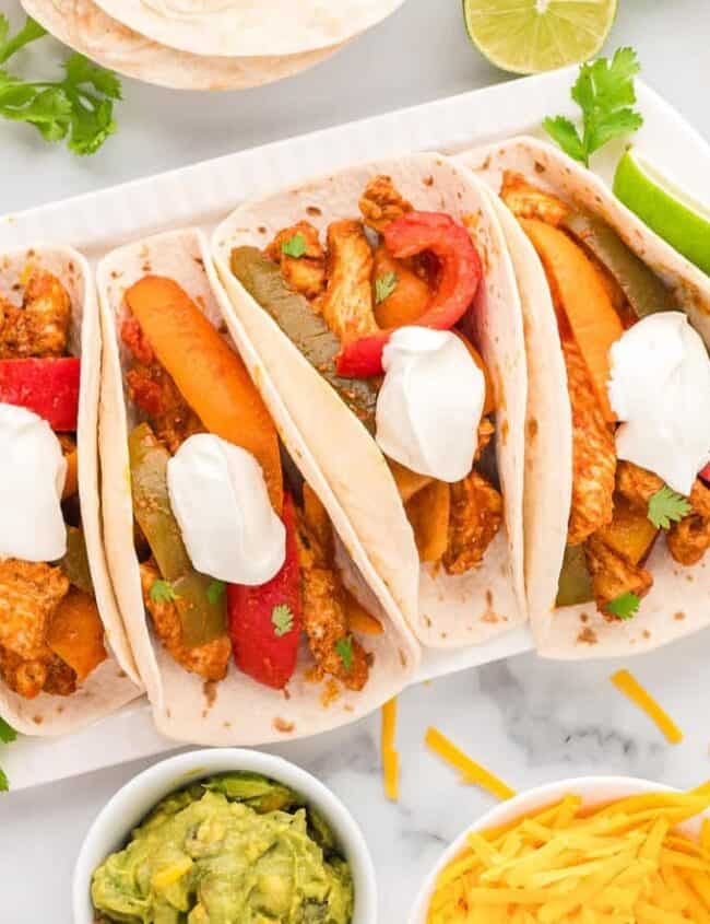 A slow cooker recipe brings these colorful tacos to life, filled with seasoned chicken, bell peppers, and onions, all topped with sour cream. Surrounding the plate are bowls of shredded cheese, guacamole, lime halves, and more tacos on a decorative napkin.