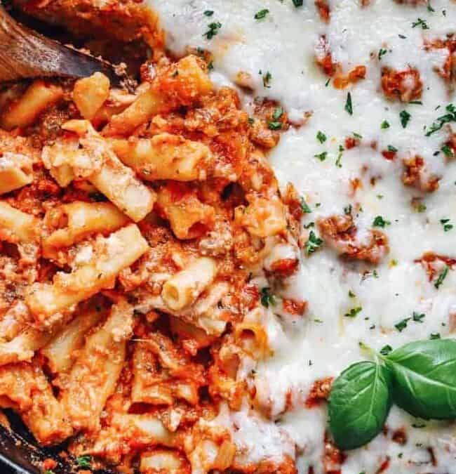 Close-up of baked ziti in a pan, with one side showing melted cheese and the other revealing pasta mixed with tomato sauce and ground meat. A wooden spoon rests in the pan, and fresh basil leaves garnish the dish.