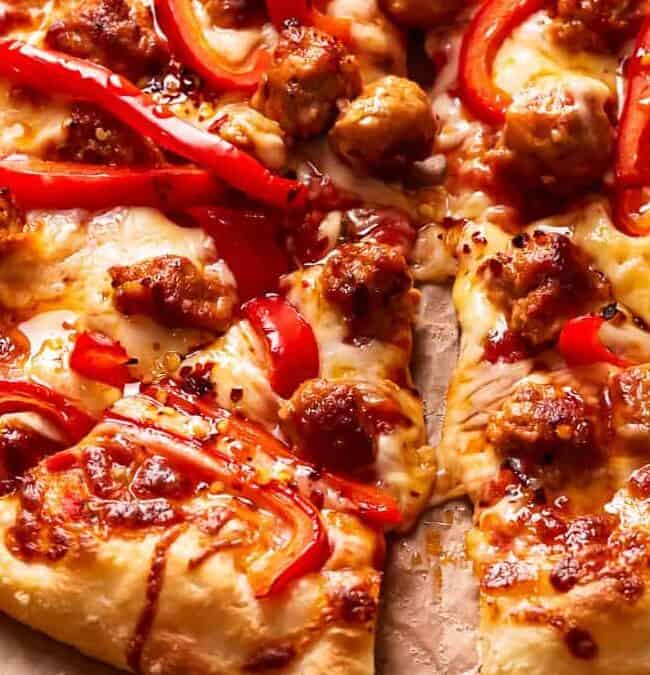 A close-up of a pizza highlighting melted cheese, sliced red bell peppers, and pieces of sausage on a wooden surface. The golden, crispy crust reflects traditional Italian recipes, with visible drops of oil enhancing its appetizing appearance.