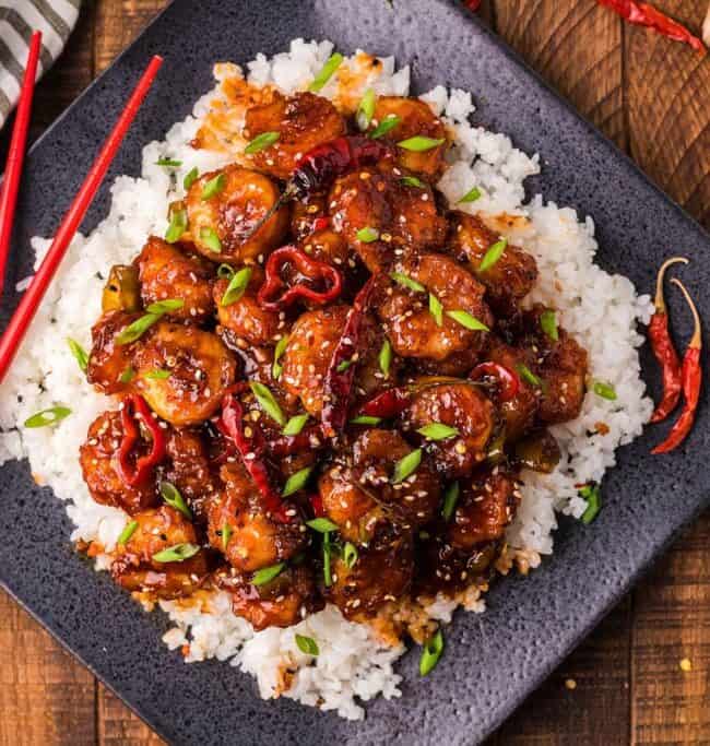 A square black plate showcases a delicious Asian recipe, featuring rice topped with glazed stir-fried meat and vibrant red and green peppers. A pair of red chopsticks rests on the side, surrounded by scattered dried chilies and a striped napkin for an authentic touch.