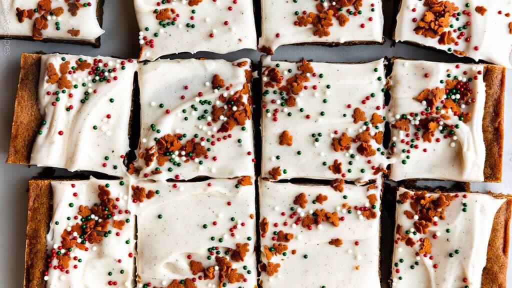 A close-up of frosted gingerbread bars cut into squares. The bars are topped with white icing and decorated with green, red, and white sprinkles, along with small gingerbread crumbs.