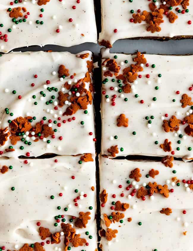 A close-up of frosted gingerbread bars cut into squares. The bars are topped with white icing and decorated with green, red, and white sprinkles, along with small gingerbread crumbs.