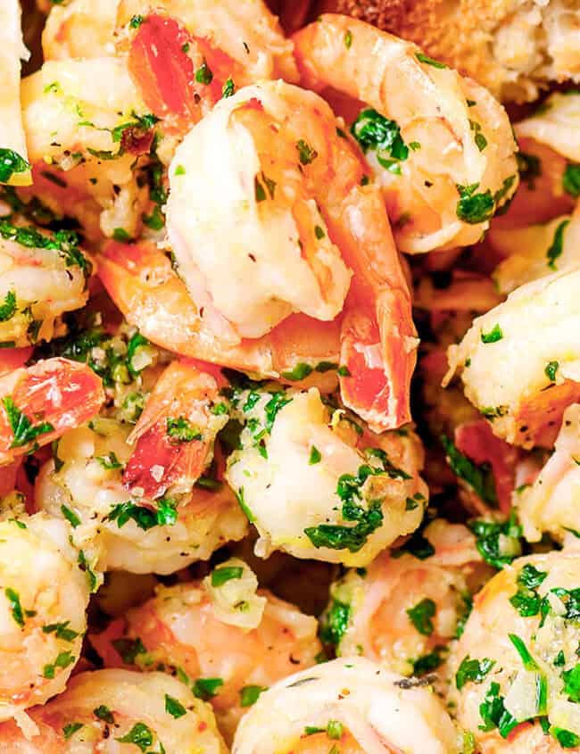 A bowl of shrimp garnished with fresh parsley and a slice of lemon. The shrimp appear cooked, with a pinkish hue, and are mixed with herbs and seasonings. A slice of crusty bread is visible at the edge.