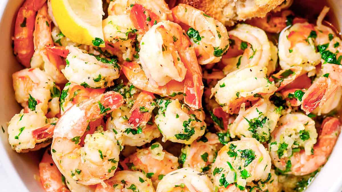 A bowl of shrimp garnished with fresh parsley and a slice of lemon. The shrimp appear cooked, with a pinkish hue, and are mixed with herbs and seasonings. A slice of crusty bread is visible at the edge.