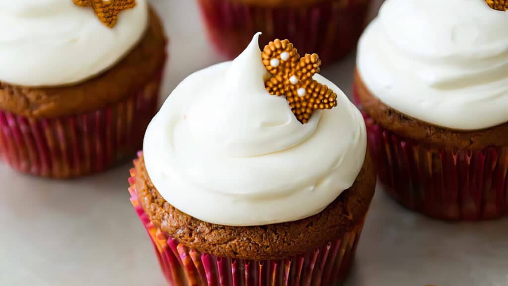 A chocolate cupcake with a swirl of white frosting, topped with a small decorative embellishment resembling a pine cone, is presented in a red cupcake liner. Other similarly decorated cupcakes are partially visible in the background.