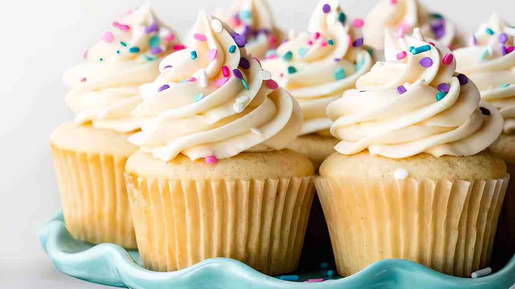 A plate of tasty vanilla cupcakes topped with swirls of creamy frosting and colorful sprinkles. Perfect for any gathering, these delightful treats are arranged closely together on a decorative, light blue platter.