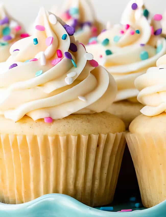 A plate of tasty vanilla cupcakes topped with swirls of creamy frosting and colorful sprinkles. Perfect for any gathering, these delightful treats are arranged closely together on a decorative, light blue platter.