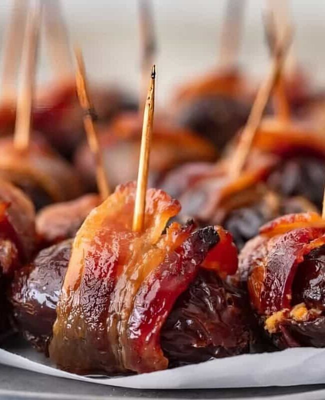 Close-up of bacon-wrapped dates on a plate. Each date is secured with a toothpick, showcasing the crispy texture of the bacon and the dark, glossy surface of the dates. The serving is arranged on parchment paper.