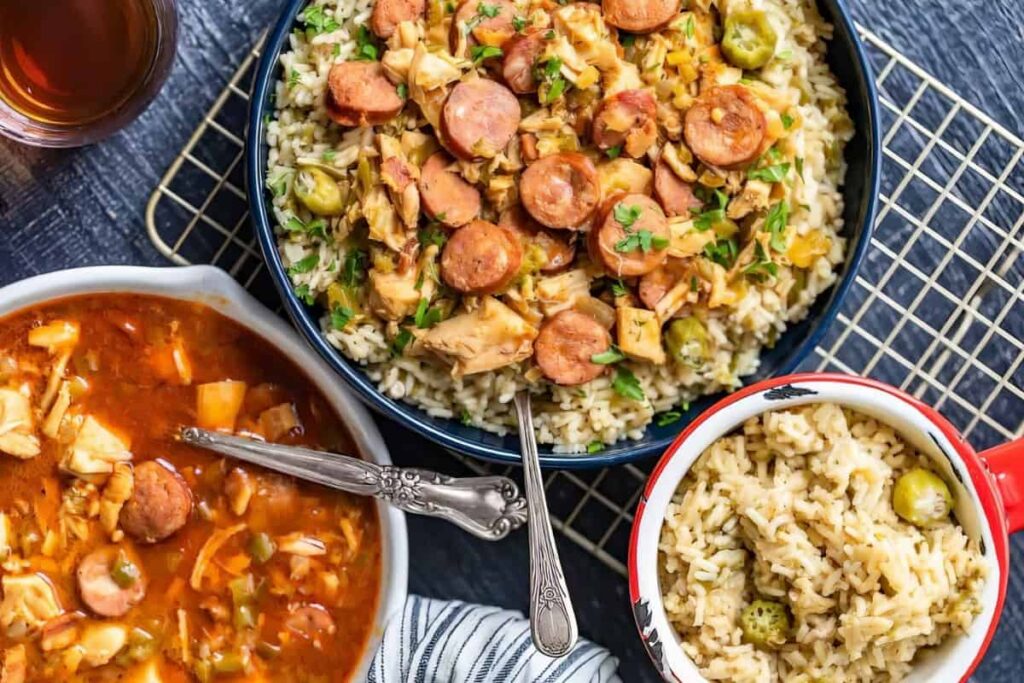 A table setting featuring a bowl of rice topped with sliced sausage and vegetables, a serving of rice in a smaller bowl, and a pot of soup with sausage and vegetables. A glass of iced tea is on the side.