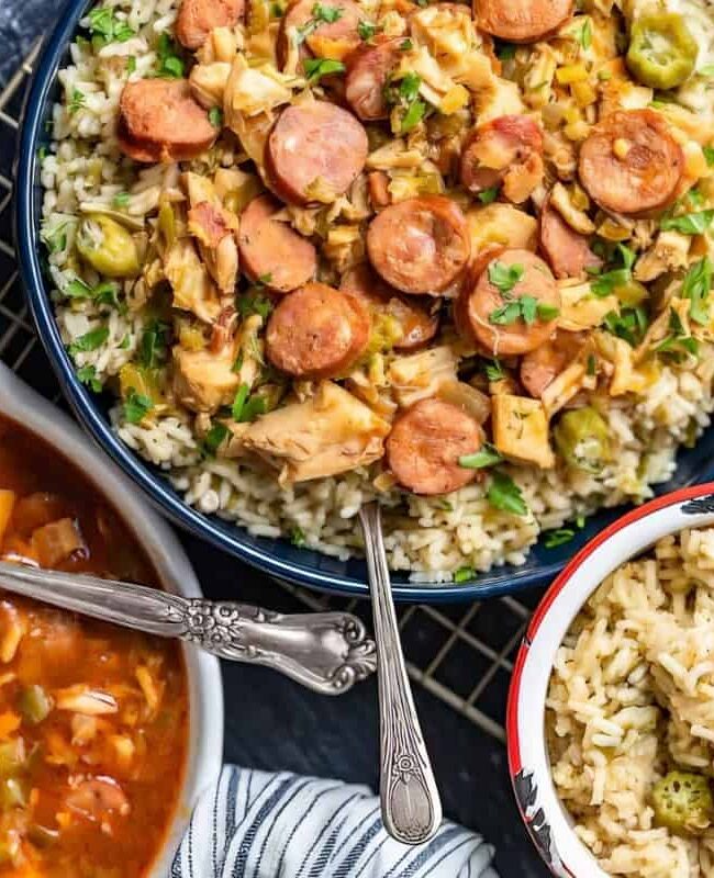 A table setting featuring a bowl of rice topped with sliced sausage and vegetables, a serving of rice in a smaller bowl, and a pot of soup with sausage and vegetables. A glass of iced tea is on the side.