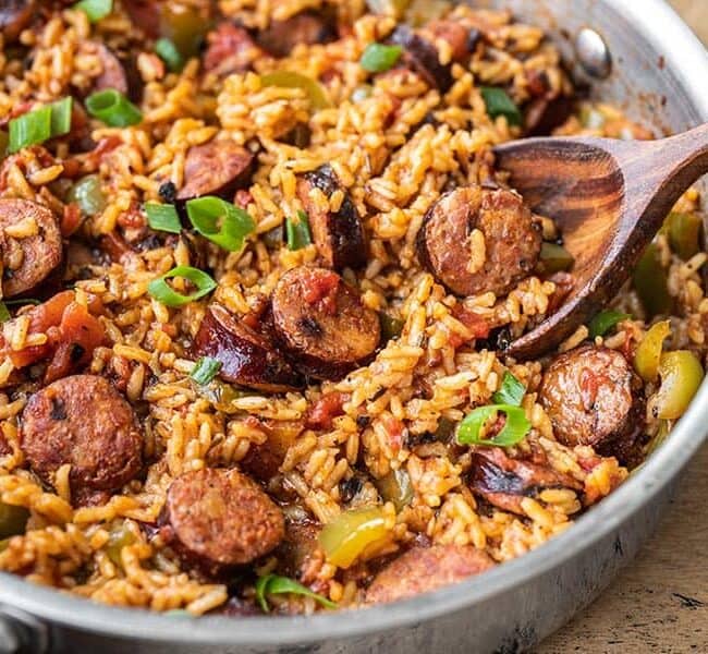 A skillet filled with Cajun jambalaya, featuring slices of sausage, rice, diced tomatoes, and green peppers. Garnished with chopped green onions, the dish appears vibrant and savory. Try this classic recipe with a wooden spoon resting invitingly in the pan.