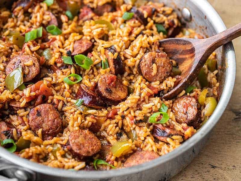 A skillet filled with Cajun jambalaya, featuring slices of sausage, rice, diced tomatoes, and green peppers. Garnished with chopped green onions, the dish appears vibrant and savory. Try this classic recipe with a wooden spoon resting invitingly in the pan.