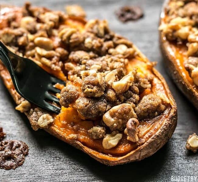 A roasted sweet potato half topped with a crunchy walnut and brown sugar crumble, on a wooden surface. A black fork is cutting into the potato.