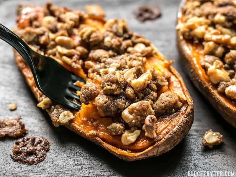 A roasted sweet potato half topped with a crunchy walnut and brown sugar crumble, on a wooden surface. A black fork is cutting into the potato.