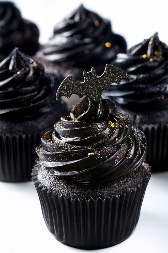 Black velvet cupcakes with glossy frosting are topped with black bat decorations and small gold specks, creating a spooky, elegant appearance. The cupcakes are arranged in a cluster on a white background.