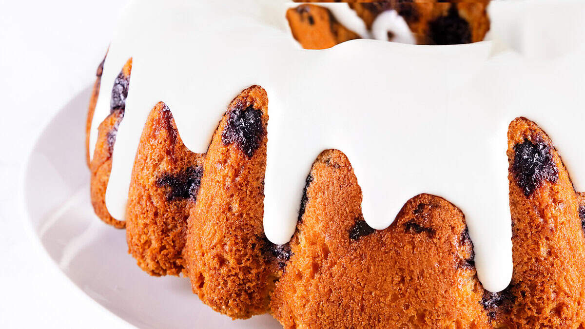 Close-up of a Bundt cake drizzled with white glaze. The cake is golden brown with visible blueberries embedded within. It is set on a white plate.