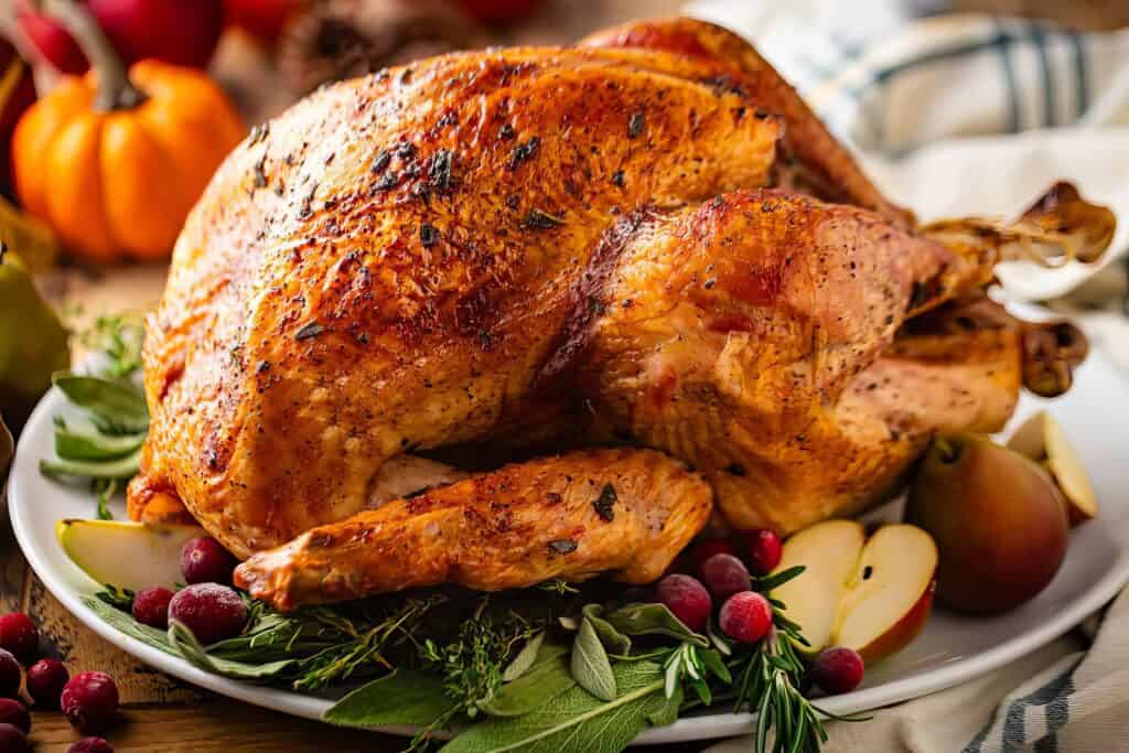 A roasted turkey on a platter surrounded by fresh herbs, apple slices, cranberries, and a small pumpkin. The turkey is golden brown and garnished, suggesting a festive or holiday meal setting.