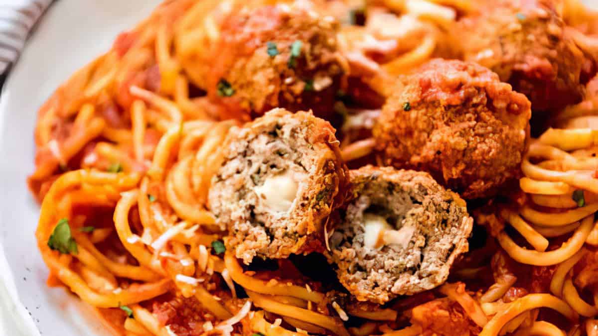 A plate of spaghetti topped with tomato sauce and meatballs. One meatball is cut in half, showing a melted cheese center. Garnished with grated cheese and parsley.