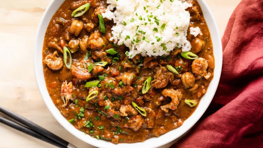 A bowl of Southern crawfish étouffée topped with white rice and garnished with chopped green onions and parsley. A red cloth and a pair of chopsticks rest beside the bowl on a light wooden surface.