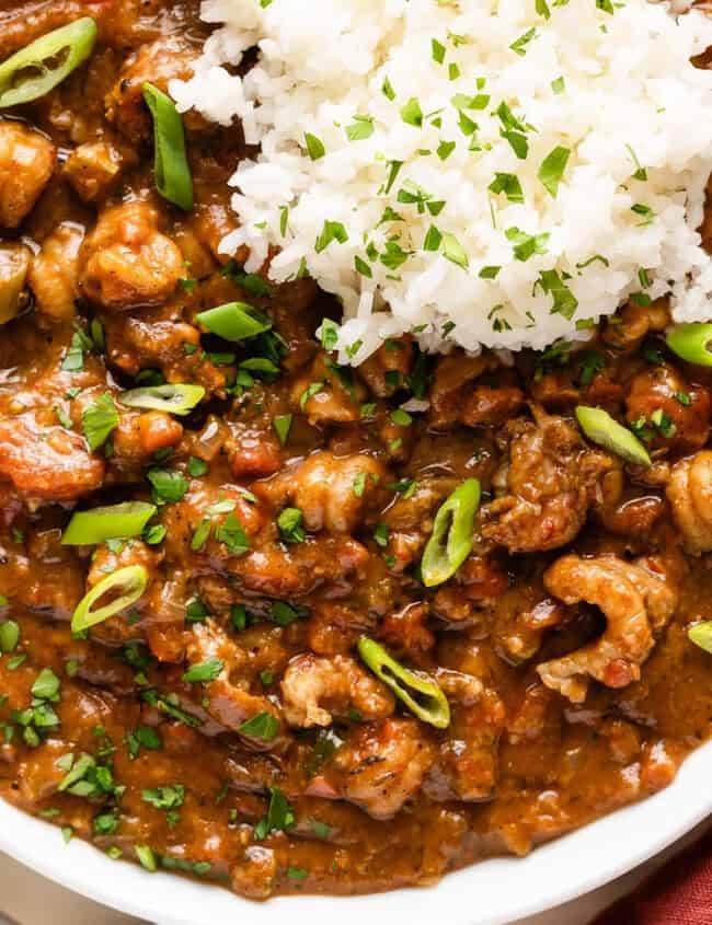 A bowl of Southern crawfish étouffée topped with white rice and garnished with chopped green onions and parsley. A red cloth and a pair of chopsticks rest beside the bowl on a light wooden surface.