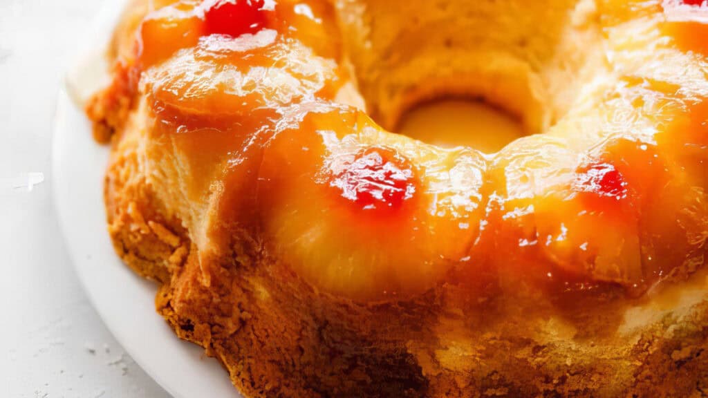 A close-up of a pineapple upside-down bundt cake on a white plate. This dessert boasts a golden brown hue with a glossy top, featuring pineapple rings and maraschino cherries beautifully embedded in a caramelized coating, showcasing the art of baking.