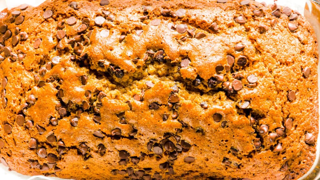 A close-up of a freshly baked banana bread topped with chocolate chips. The bread has a golden-brown crust with a slightly cracked surface, and it fills the frame with its texture and rich color.