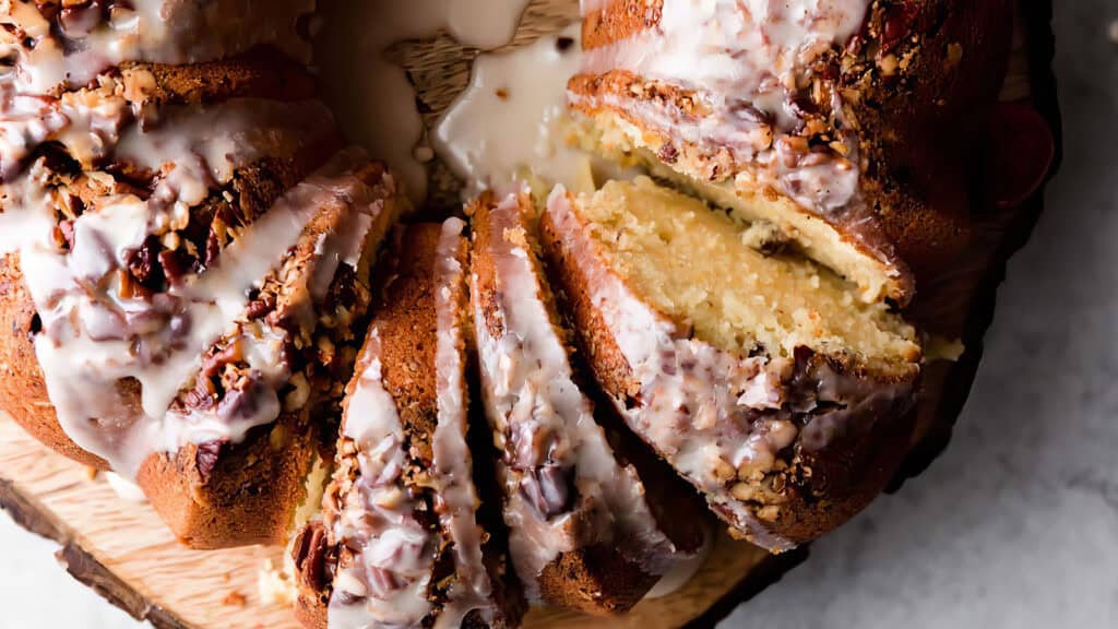 A luscious bundt cake with chopped nuts is elegantly sliced on a wooden platter. Its creamy interior and drizzled icing are prominently visible, inviting everyone to savor this delightful treat.