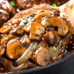 Close-up of a savory Salisbury Steak covered in a rich mushroom and onion gravy, served in a cast-iron skillet. The dish is garnished with fresh herbs, and the sauce glistens, highlighting the tender meat's juicy texture.