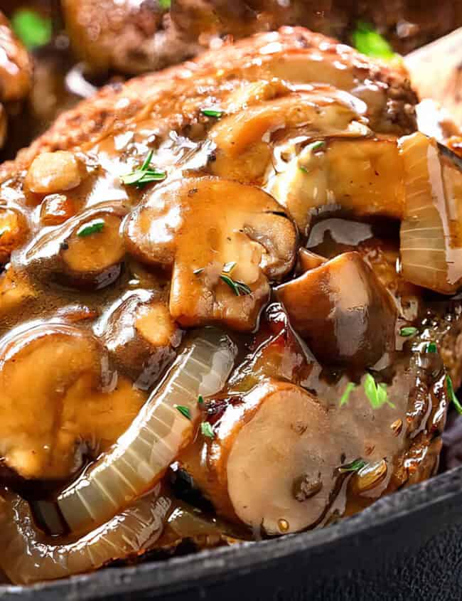 Close-up of a savory Salisbury Steak covered in a rich mushroom and onion gravy, served in a cast-iron skillet. The dish is garnished with fresh herbs, and the sauce glistens, highlighting the tender meat's juicy texture.