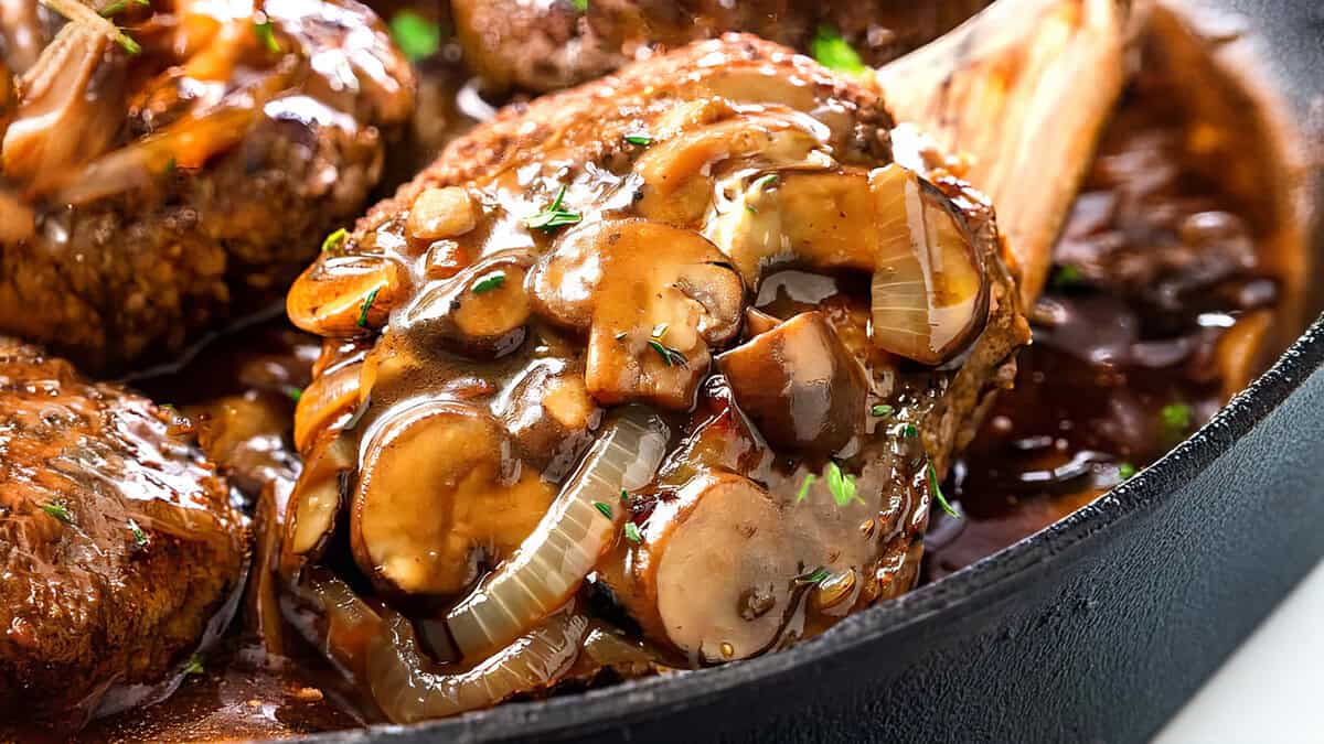 Close-up of a savory Salisbury Steak covered in a rich mushroom and onion gravy, served in a cast-iron skillet. The dish is garnished with fresh herbs, and the sauce glistens, highlighting the tender meat's juicy texture.