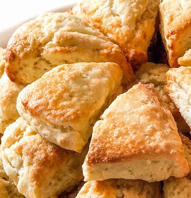 A heap of freshly baked scones piled on a white plate. The scones are golden brown and have a fluffy texture, suggesting they are warm and ready to eat.