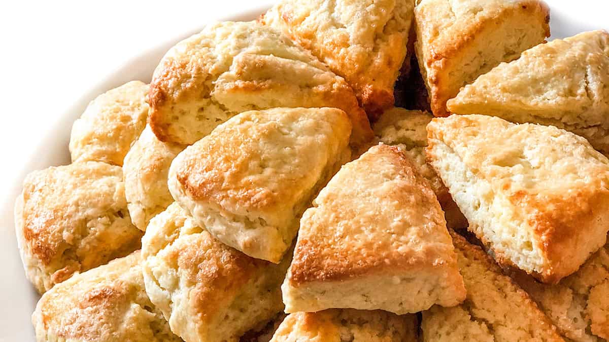 A heap of freshly baked scones piled on a white plate. The scones are golden brown and have a fluffy texture, suggesting they are warm and ready to eat.