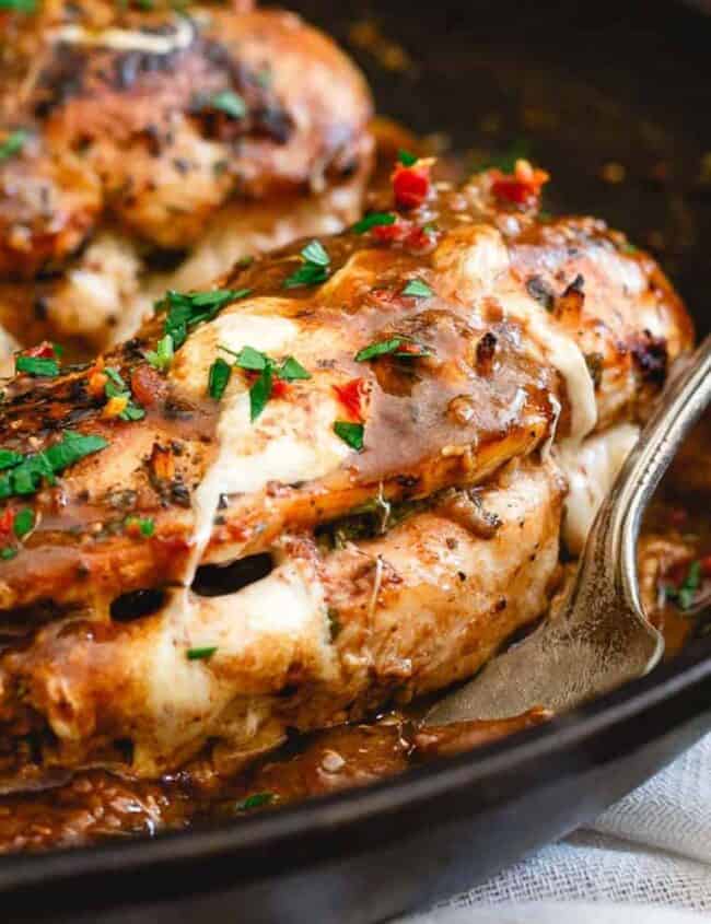 A close-up of a skillet features seasoned, cooked chicken breasts stuffed with herbs and melted cheese, embodying the essence of delectable chicken recipes. The dish is garnished with chopped parsley, with a spoon resting beside the chicken, and scattered parsley leaves around the skillet.