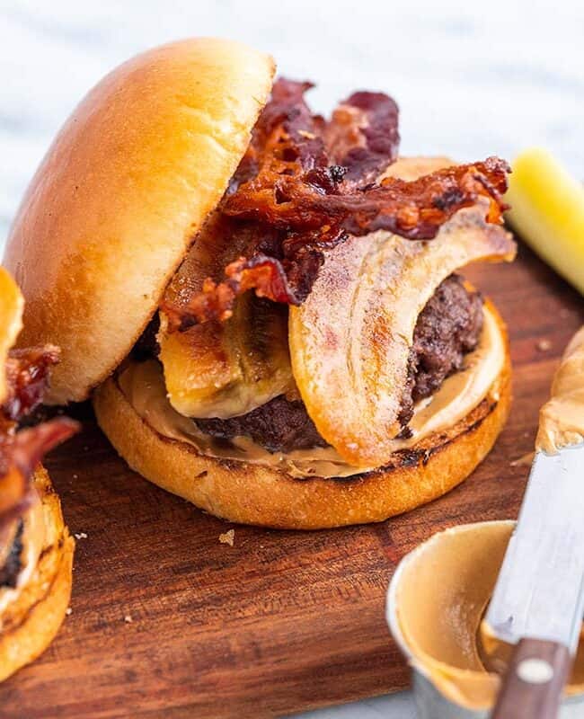 Two juicy burgers featuring crispy bacon and creamy peanut butter sit enticingly on a wooden board, accompanied by sliced pickles and a knife generously coated with peanut butter. This creative twist on classic bacon recipes is set against an elegant marble surface.