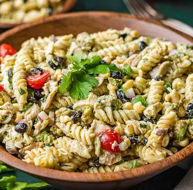 A wooden bowl brimming with a beloved pasta salad recipe: rotini, cherry tomatoes, olives, corn, and herbs, elegantly garnished with fresh parsley. This creamy and colorful delight sits on a green tablecloth with a fork poised in the background.