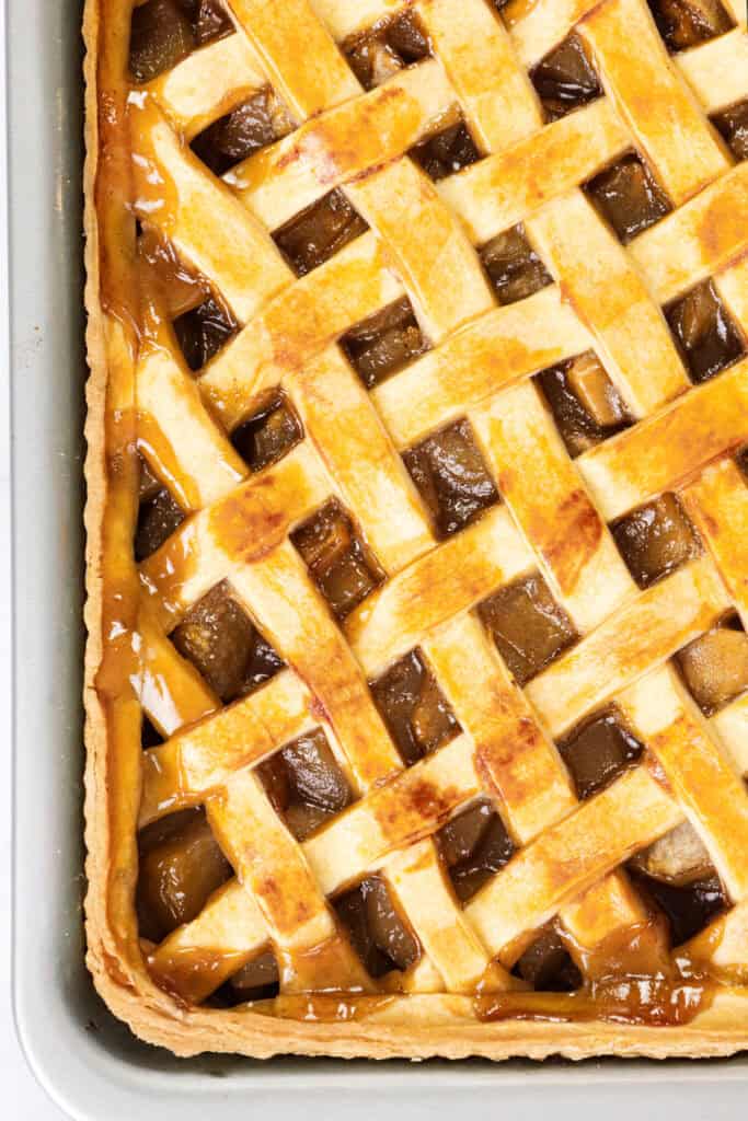 A close-up of a freshly baked pear pie with a golden-brown lattice crust. The filling, visible between the strips, appears to be a caramelized fruit mixture. The pie sits in a rectangular baking pan, its warm aroma inviting and comforting.