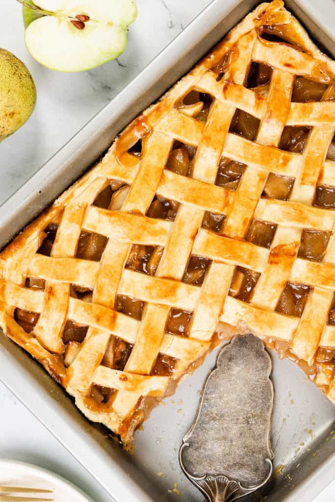 A top view of a freshly baked apple pie showcases its lattice crust in a baking dish, surrounded by sliced green apple and pear on the marble surface. A pie server rests beside the tempting creation, with one delectable slice already missing.