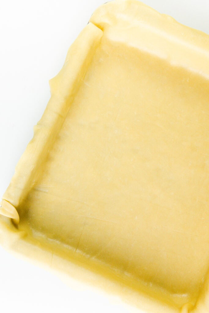 A square baking tray lined with uncooked pastry dough, ready to transform into a delightful apple pie, edges folded over the sides, set against a crisp white background.