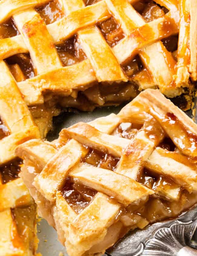 Close-up of a freshly baked pie with a golden lattice crust. A slice is being lifted, revealing the soft, cinnamon-spiced apple and pear filling. The lattice top is glossy and caramelized.