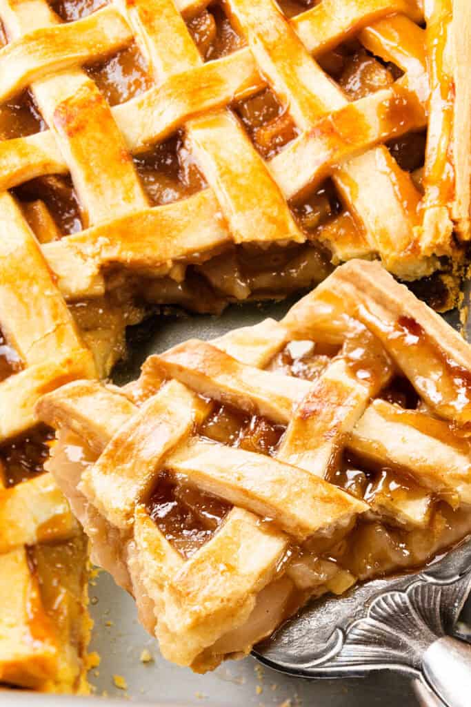 Close-up of a freshly baked pie with a golden lattice crust. A slice is being lifted, revealing the soft, cinnamon-spiced apple and pear filling. The lattice top is glossy and caramelized.