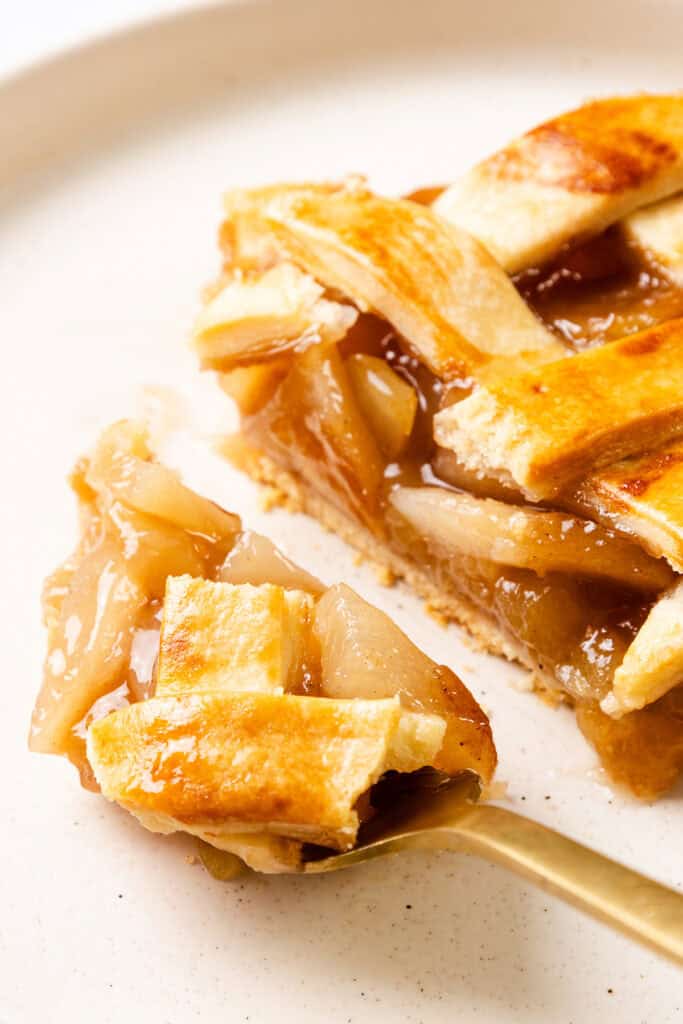 A close-up of a slice of apple pie on a white plate showcases its golden, lattice crust and gooey filling. A fork holds a small piece, highlighting the exquisite texture and layers, making this classic apple delight irresistible.